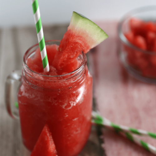 Delicious Watermelon Lemonade Slushy Recipe 14 watermelon slushing in clear glass sitting on table with green and white straw, slice of watermelon on the rim of the glass and on the side.
