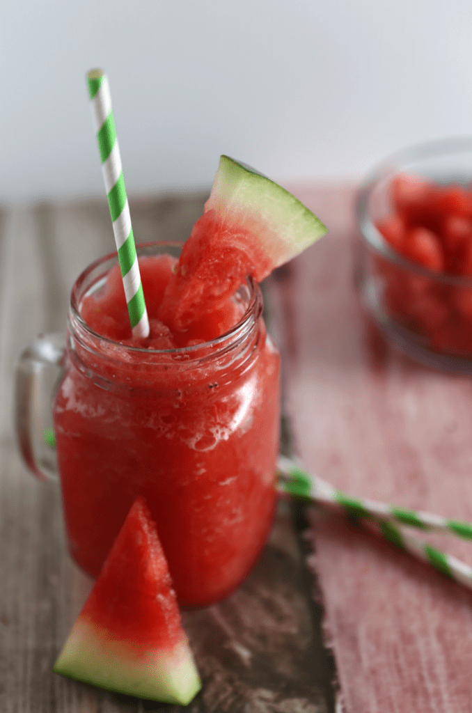 Easy Summer Recipes Roundup: Beat The Heat 16 Simple Recipes 13 watermelon slushing in clear glass sitting on table with green and white straw, slice of watermelon on the rim of the glass and on the side.