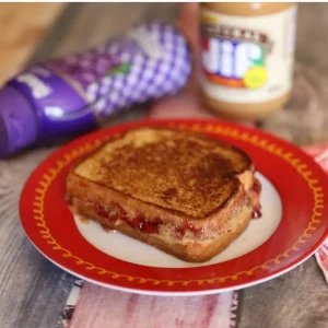 The Ultimate Grilled Peanut Butter and Jelly Sandwich Recipe 20 A perfectly toasted, golden-brown grilled peanut butter and jelly sandwich sliced in half on a red plate, showing the warm, melted Jif Natural peanut butter and gooey Smucker's grape jelly oozing from the sides.