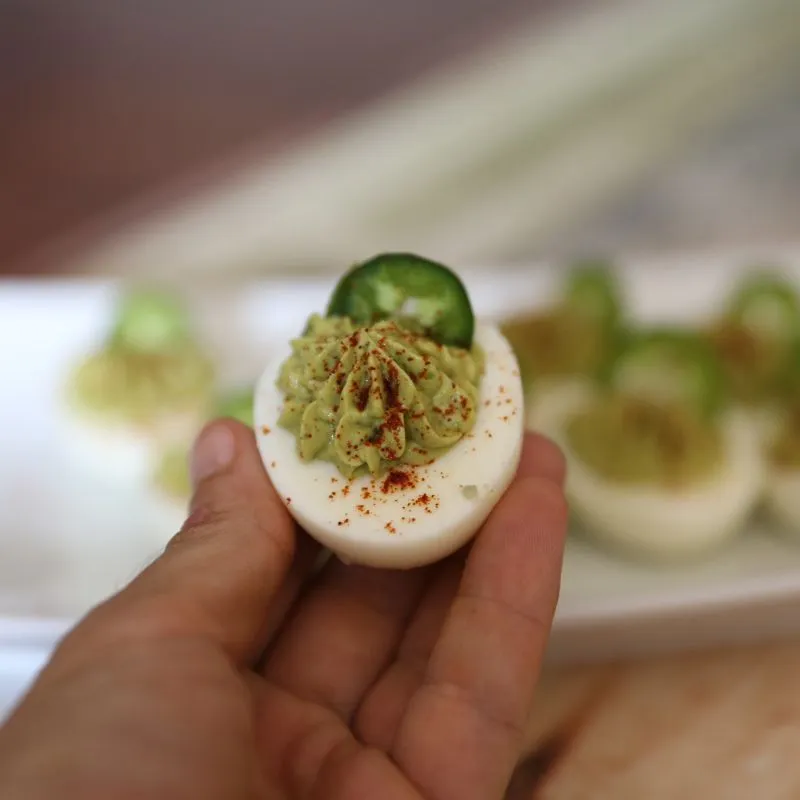 Hand holding one Holy Guacamole Deviled Egg with plateful in the background on rustic wooden table.