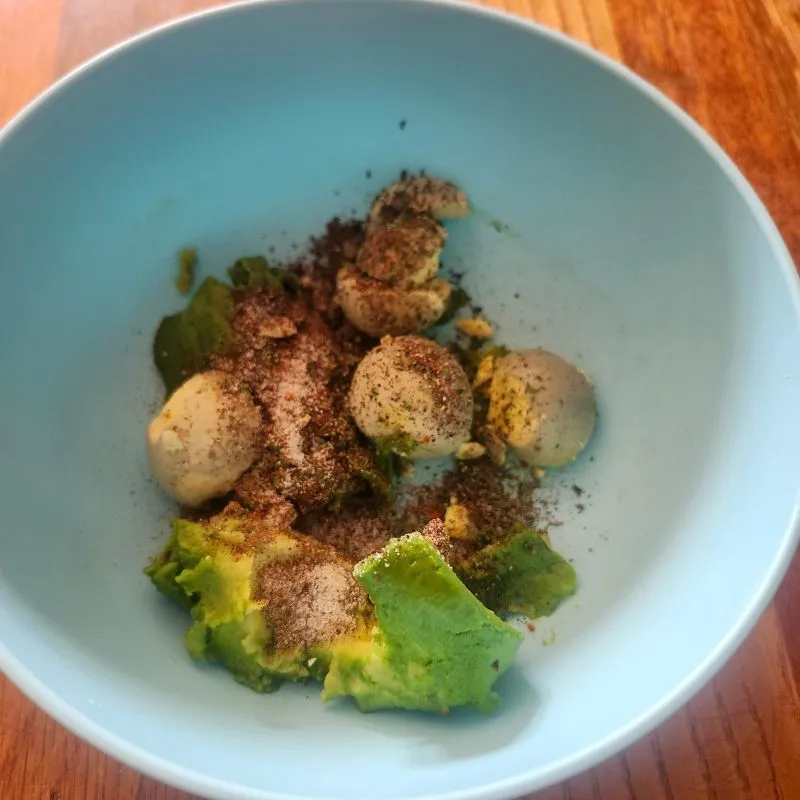 seasonings, avocado and hard boild egg yolks in a blue bowl ready to be mixed for Holy Guacamole Deviled Eggs