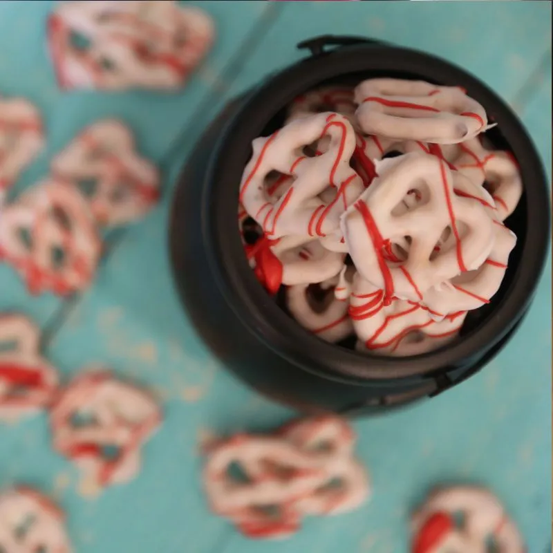 pretzels covered in white chocolate with red candy melts splattered on them to look like blood in a black caldron