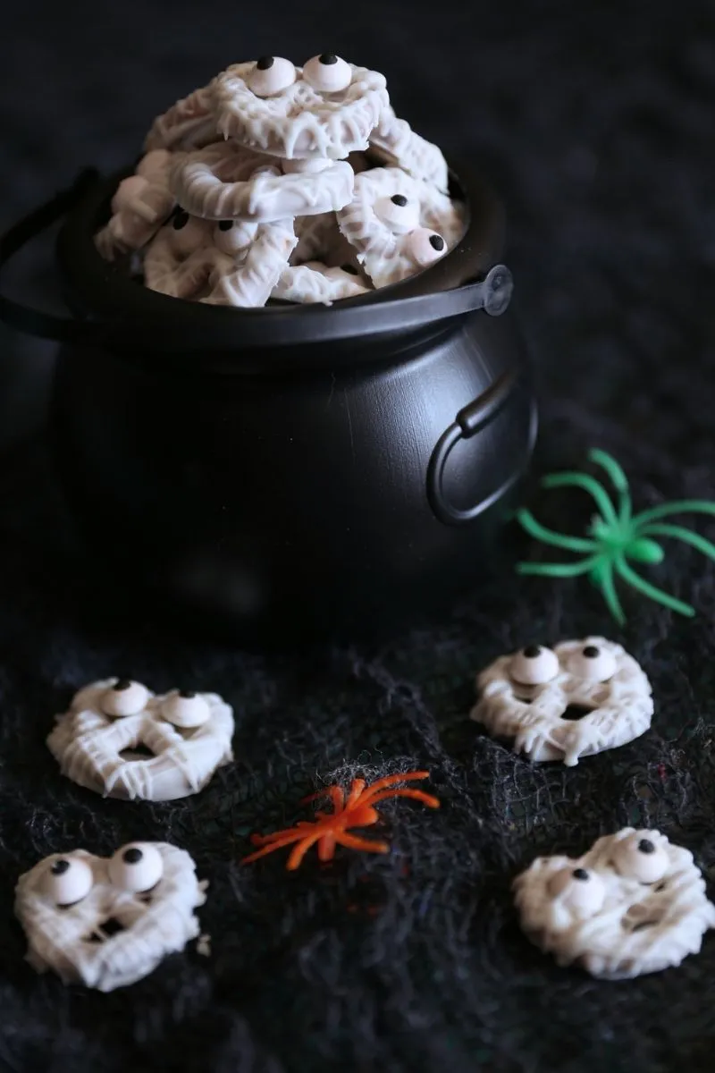A bowl of Easy Halloween White Chocolate Mummy Pretzels with candy eyes, served next to a small black cauldron and a green toy spider on a dark, spooky background.