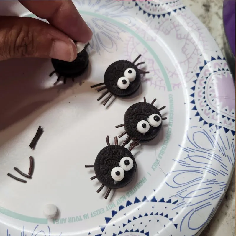 three oreo mini spiders on a paper plate and a hand placing eyes o the last spider.