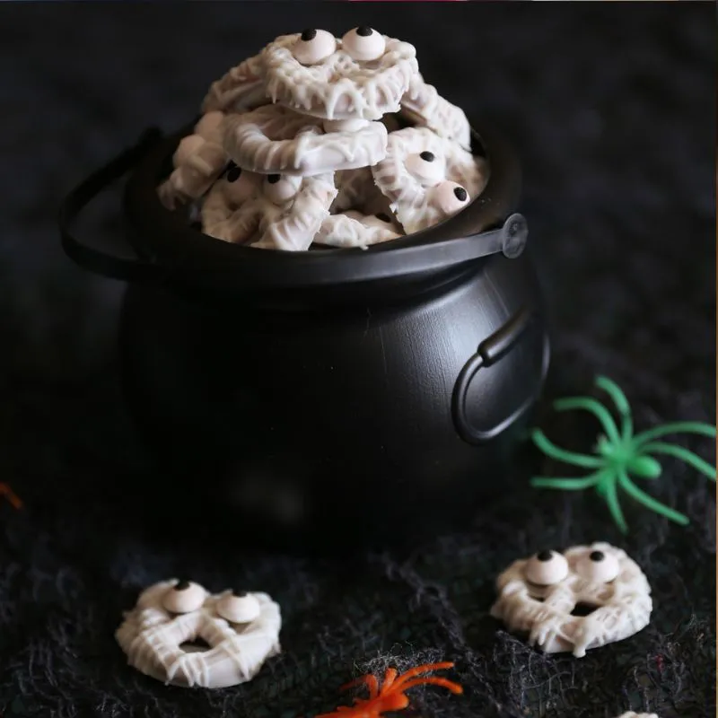 A black cauldron filled with white chocolate mummy pretzels, garnished with candy eyes and a green toy spider on a textured black background.