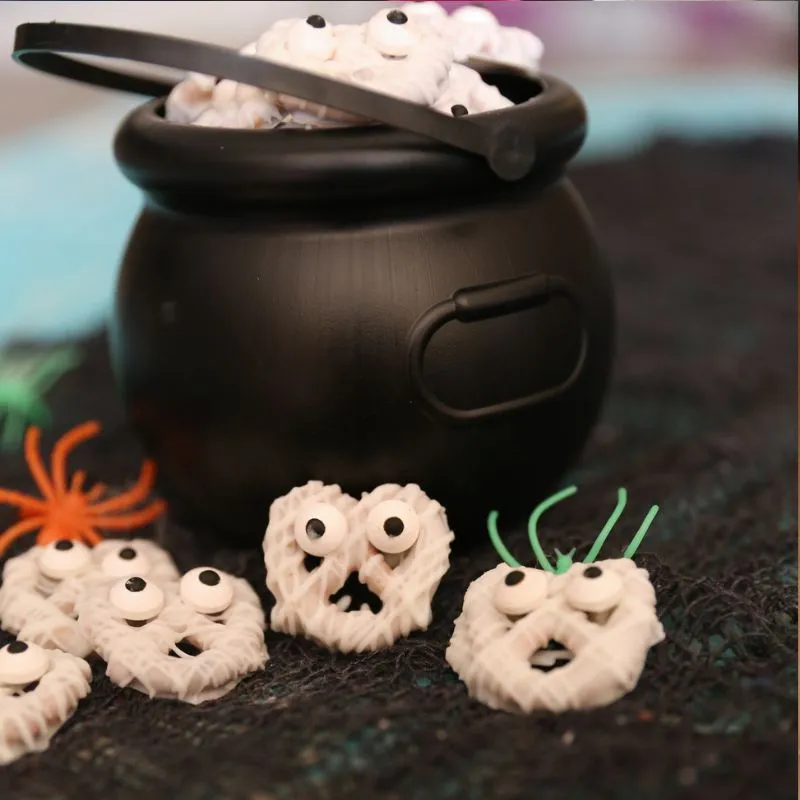 White chocolate mummy pretzels with candy eyes piled in a black cauldron on a dark, Halloween-themed surface.