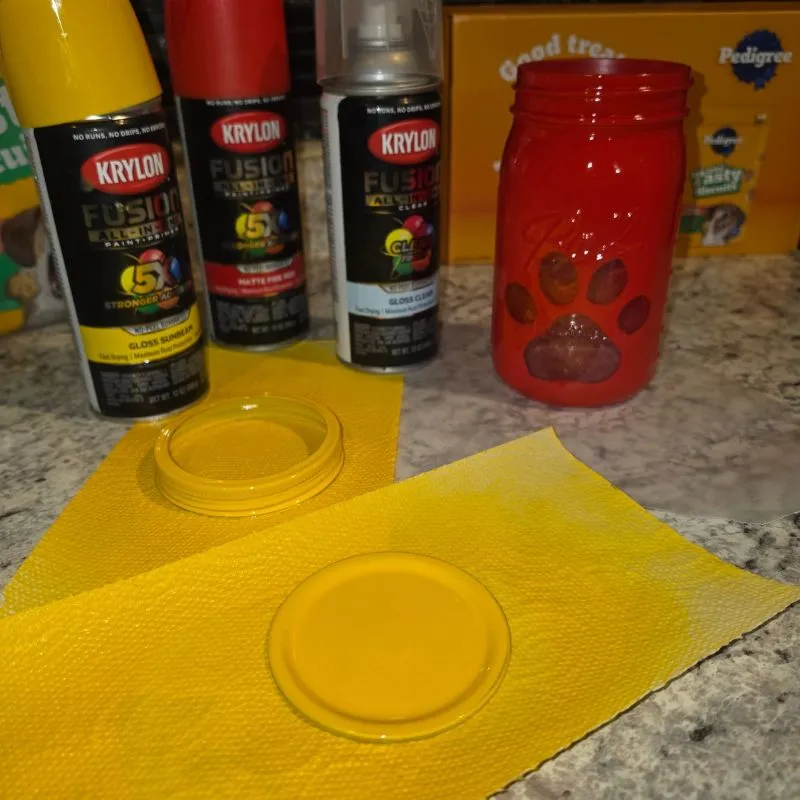 Awesome DIY Dog Treat Jar Using a Mason Jar 17 Red spray painted mason jar with homemade paw print stencil mason jar lid painted yellow next to three cans of spray paint.